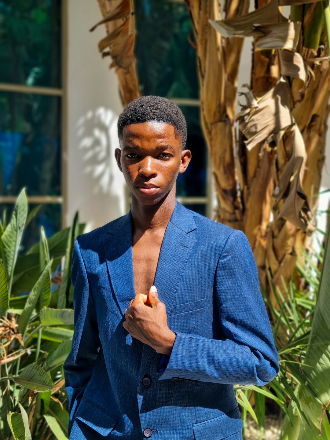 Man in a blue suit jacket stands outside near foliage.